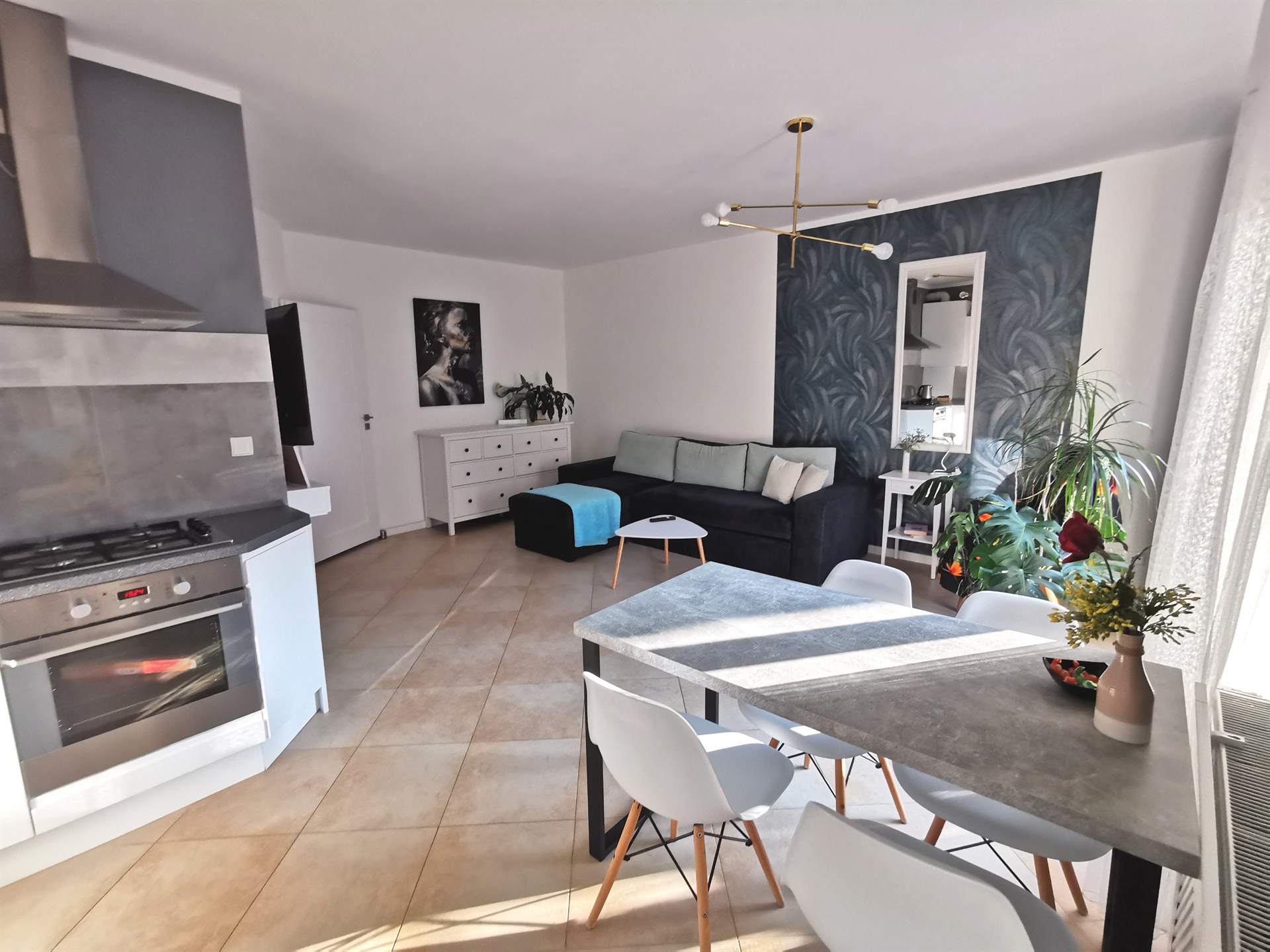 Jasne wnętrze: kuchnia z piecem, stół z białymi krzesłami i wazonem, ciemnoszara sofa, rośliny przy oknie