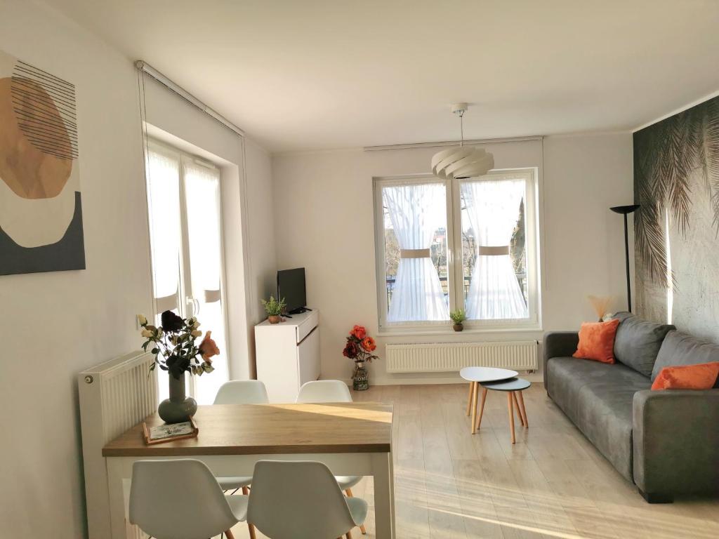 Jasny salon z szarą sofą i pomarańczowymi poduszkami, drewniany stół, dwa okna z firankami