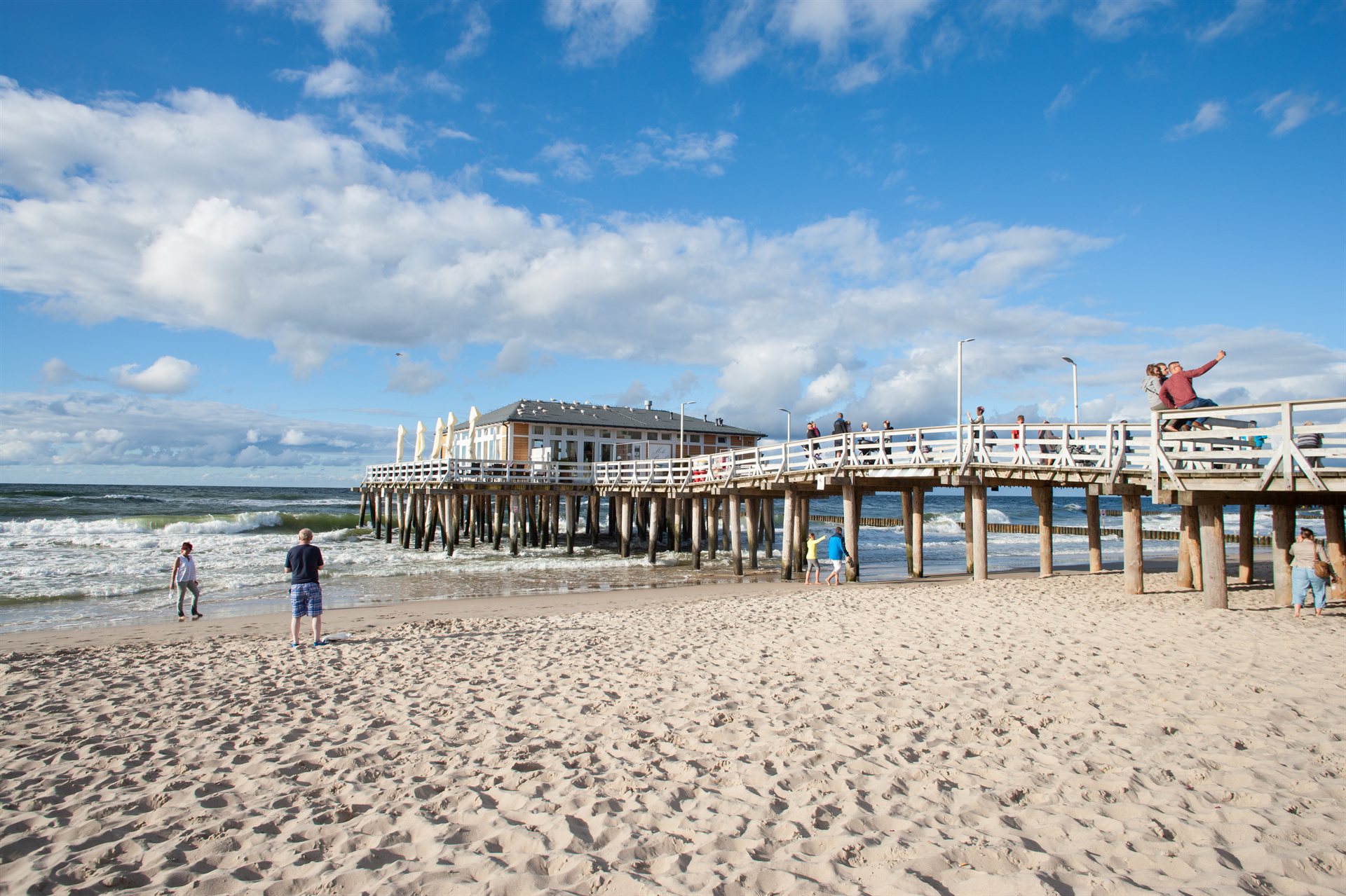 Drewniane molo z restauracją na palach nad wzburzonym morzem, piaszczysta plaża i spacerujący ludzie