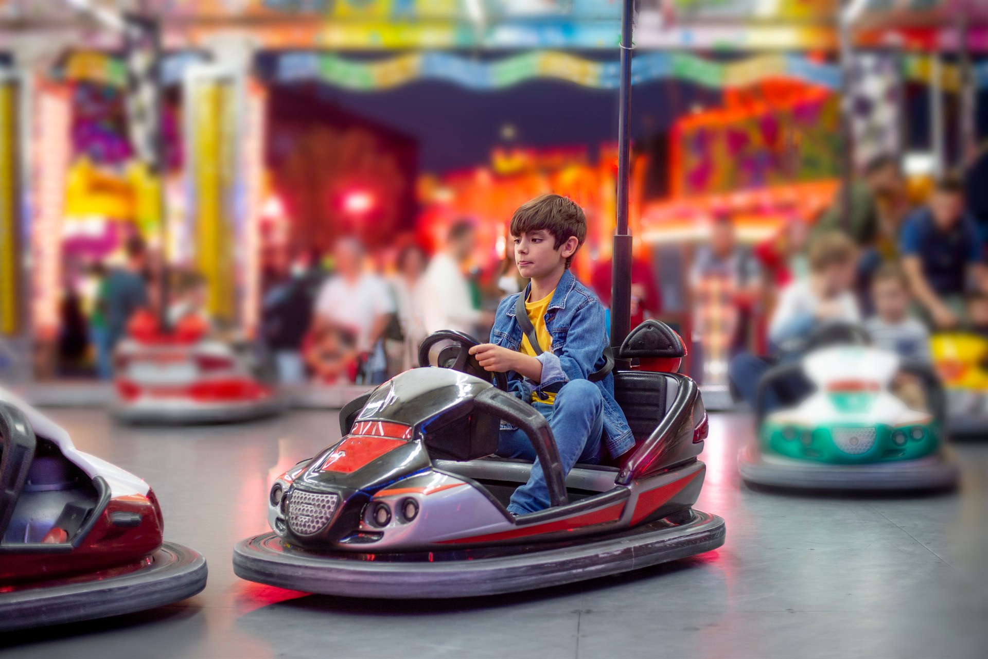 Chłopiec w dżinsowej kurtce kieruje czerwono-srebrnym samochodzikiem bumperowym przy kolorowych światłach