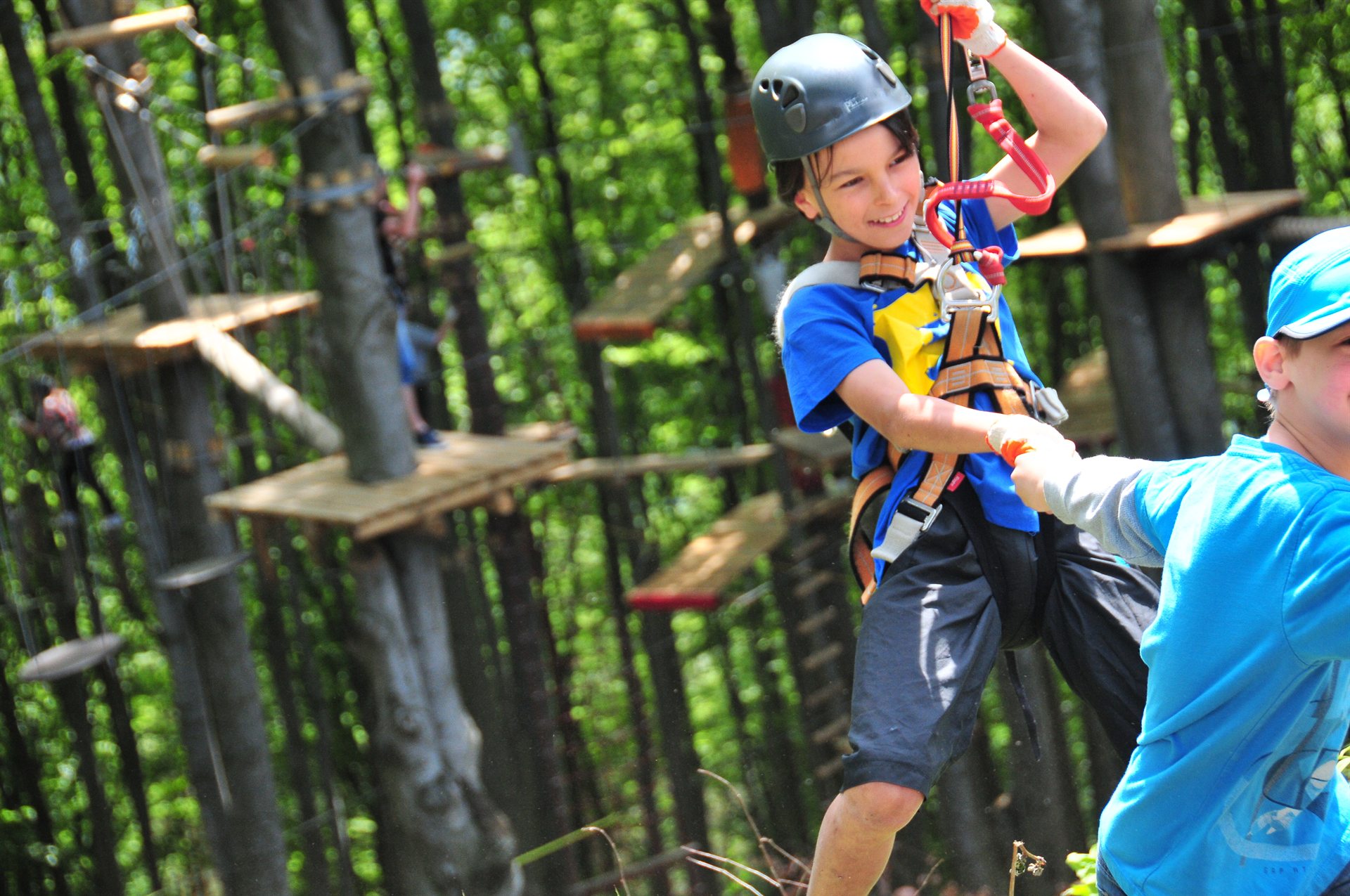 Uśmiechnięty chłopiec w kasku i uprzęży trzyma się liny na parku linowym wśród drzew
